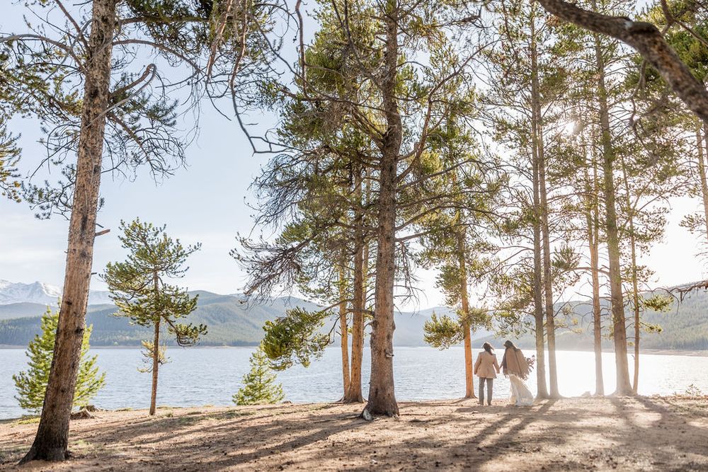 Couple walking among tall pine trees near mountain lake with sunlight streaming through branches on scenic shore.