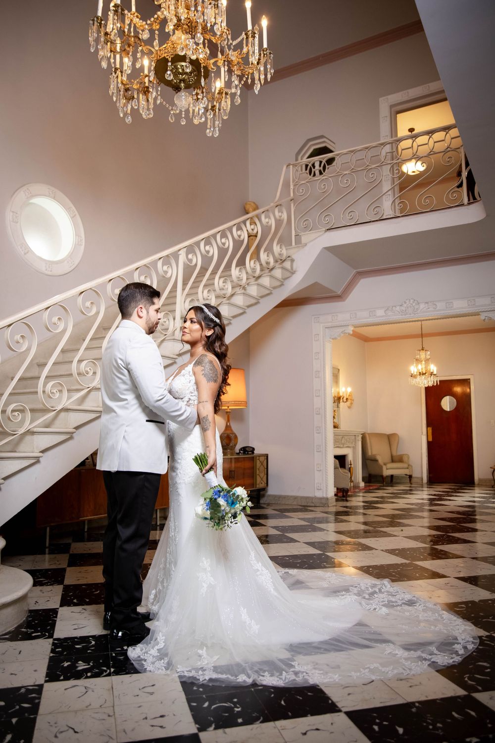 Elegant wedding couple embraces in grand foyer with crystal chandelier and white spiral staircase on checkered floor.