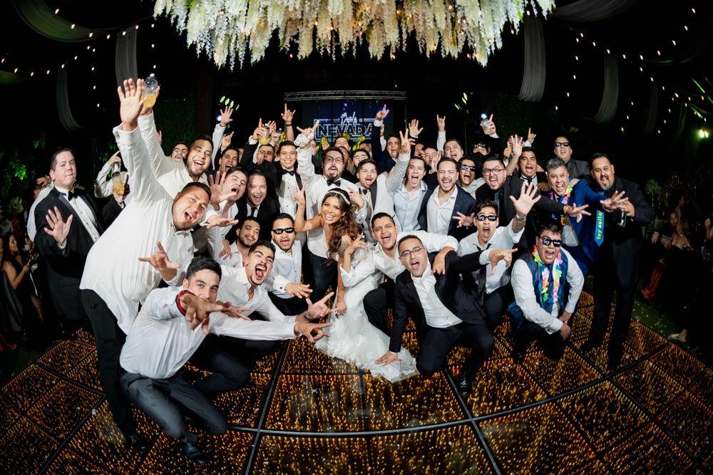 Large wedding party poses together beneath elegant chandelier lighting at reception venue.