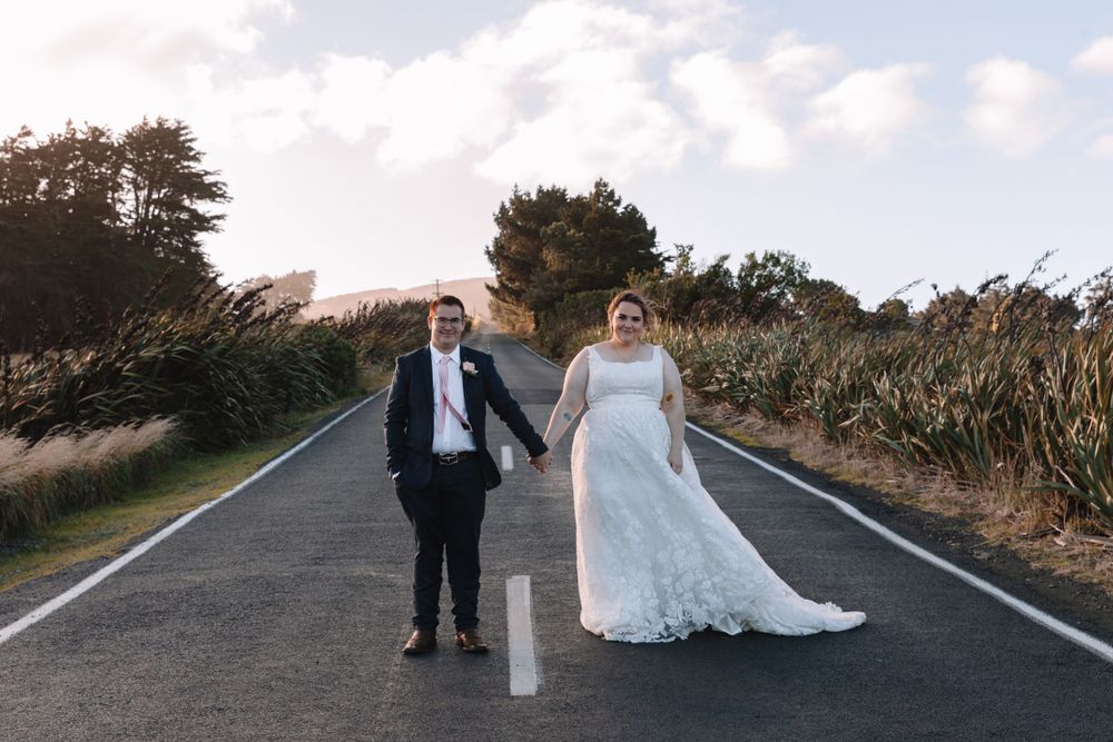 Newlyweds hold hands while standing on empty road with natural scenery.