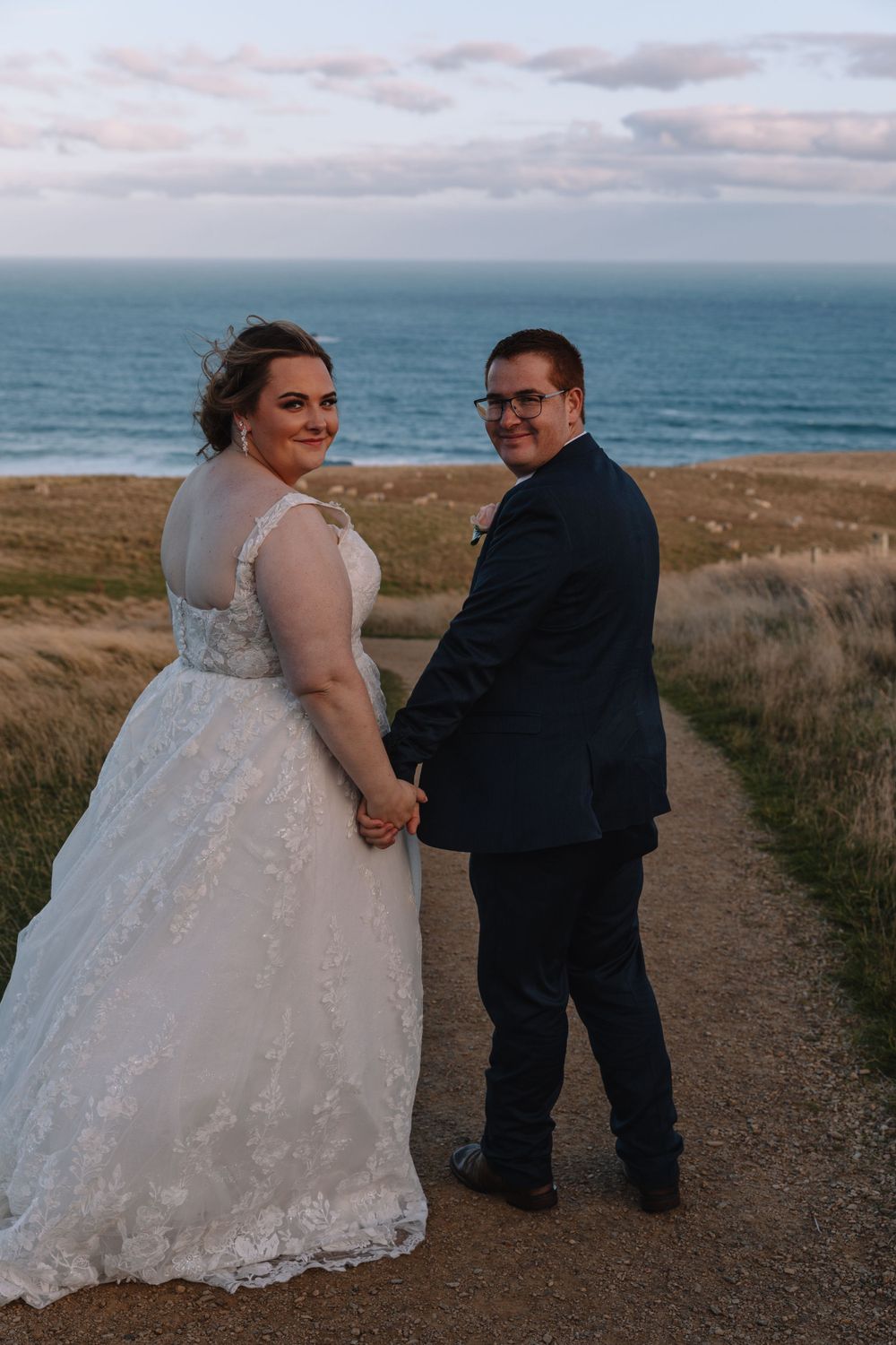 A couple in wedding attire poses on a coastal path overlooking the ocean during sunset.