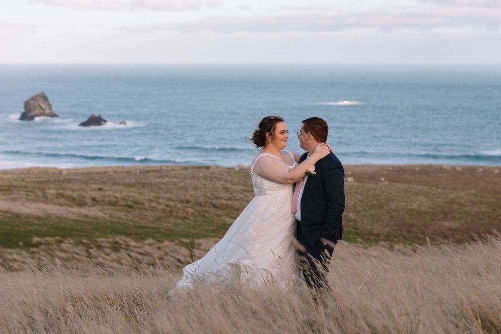 Newlyweds share a romantic moment in a grassy field with ocean rocks visible in the background.