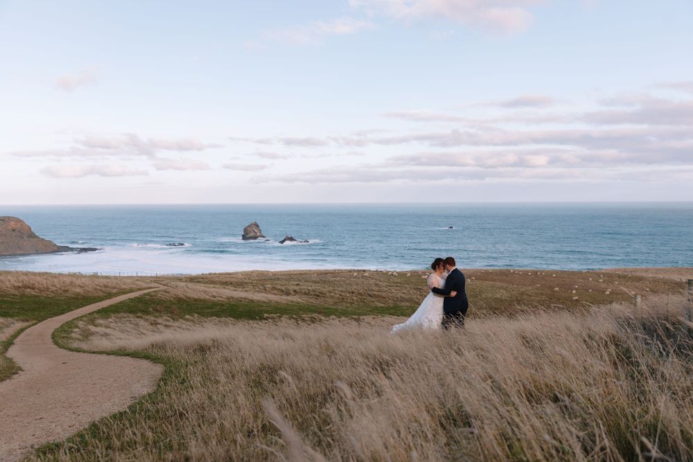 Wedding couple stands in tall grass on a winding coastal path with ocean views.