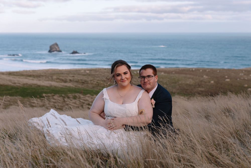Newlyweds sit together in coastal grassland with rocky ocean outcrops in the background.