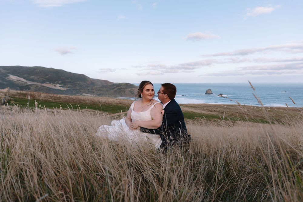 A joyful wedding moment captured in tall grass with ocean views and rocky coastline.