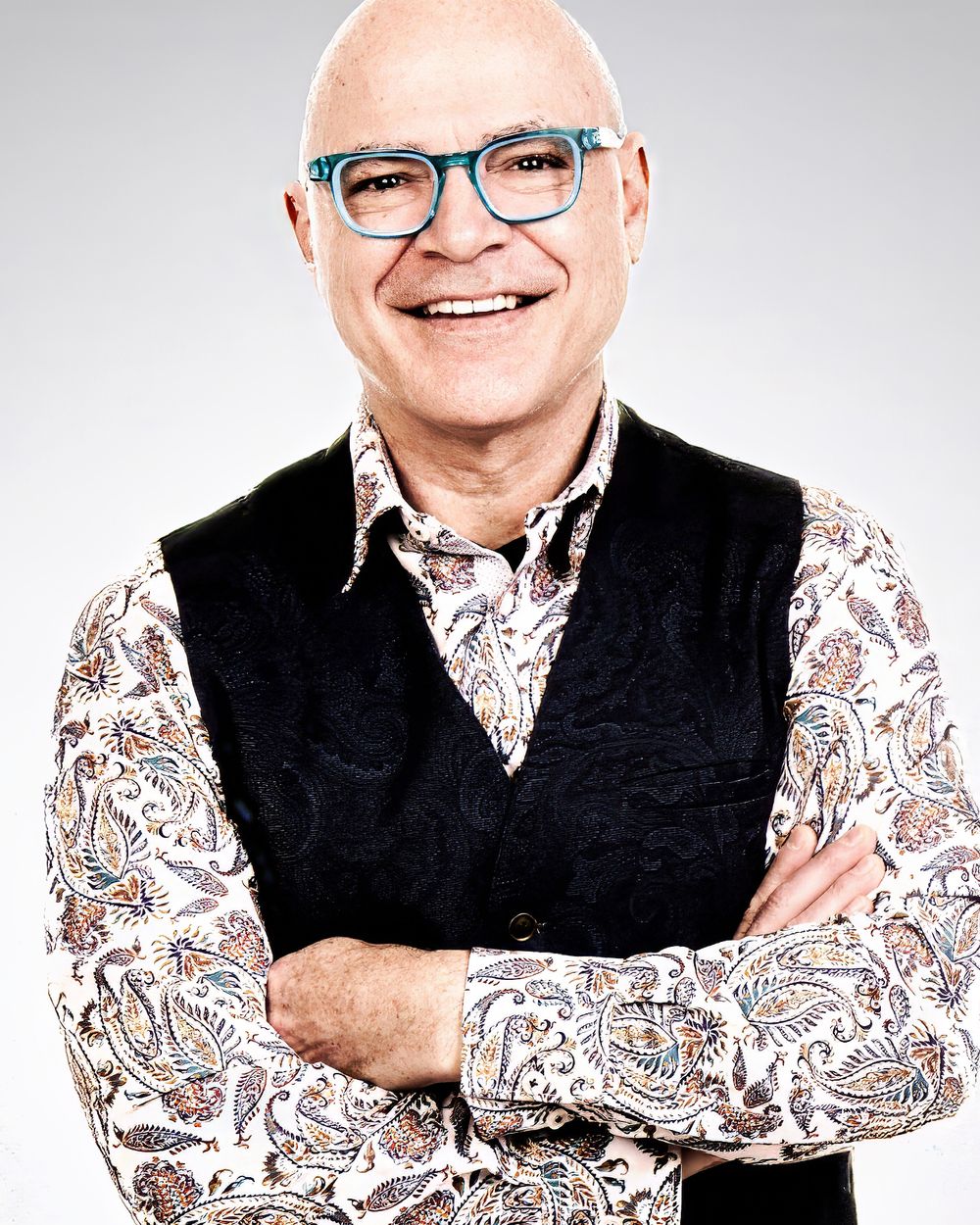 Professional portrait of someone wearing glasses, a floral print shirt and black vest, standing with arms crossed and smiling.