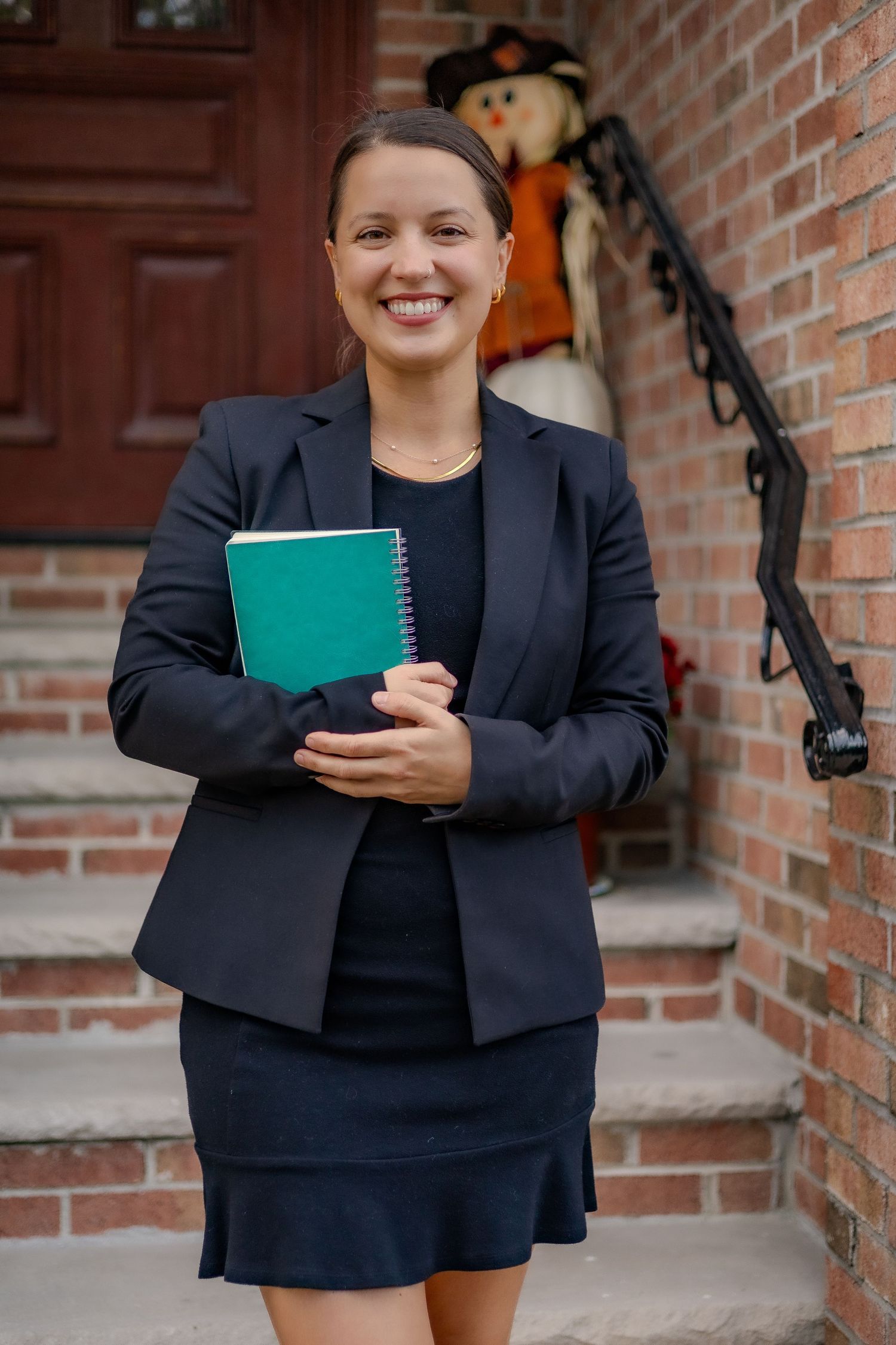 Professional in a navy business suit holds a teal notebook while standing on brick steps with autumn decor visible.