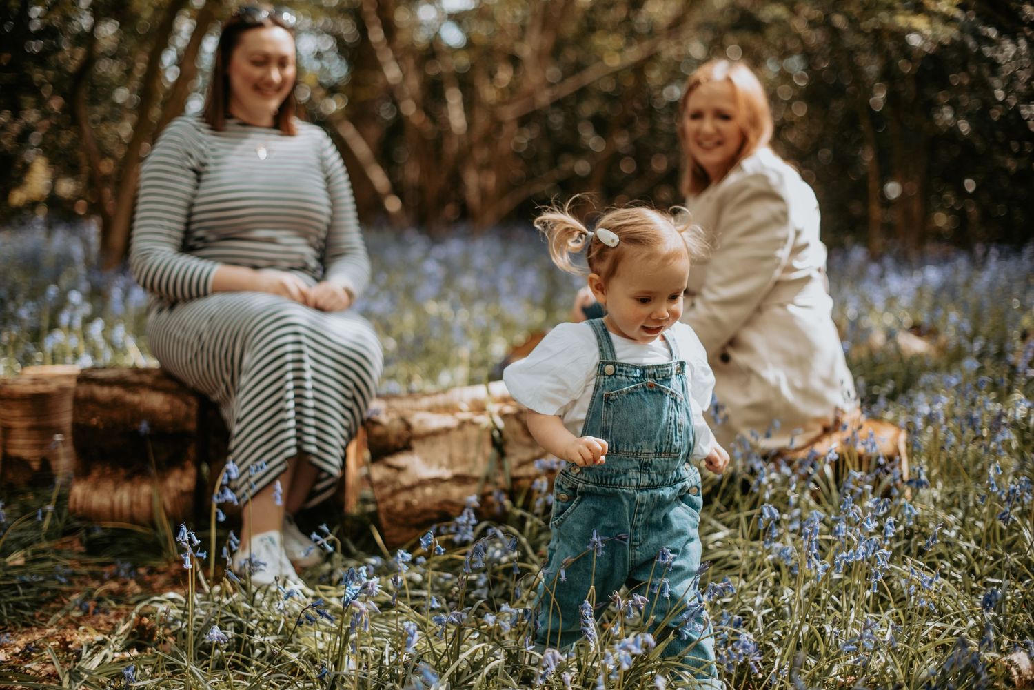 A toddler in denim overalls explores a spring bluebell garden while adults sit on a rustic log nearby.