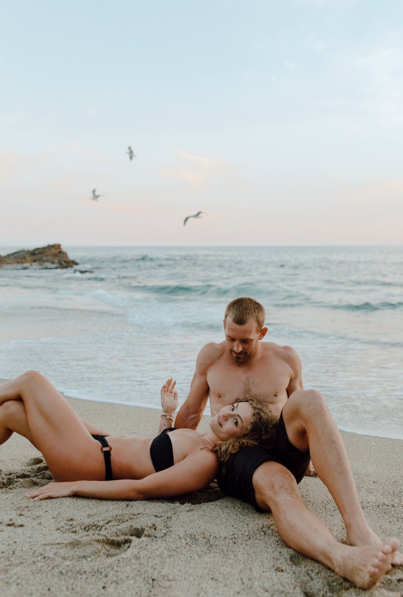 A couple shares an intimate moment together on a sandy beach at sunset with seagulls flying overhead and waves crashing nearby.