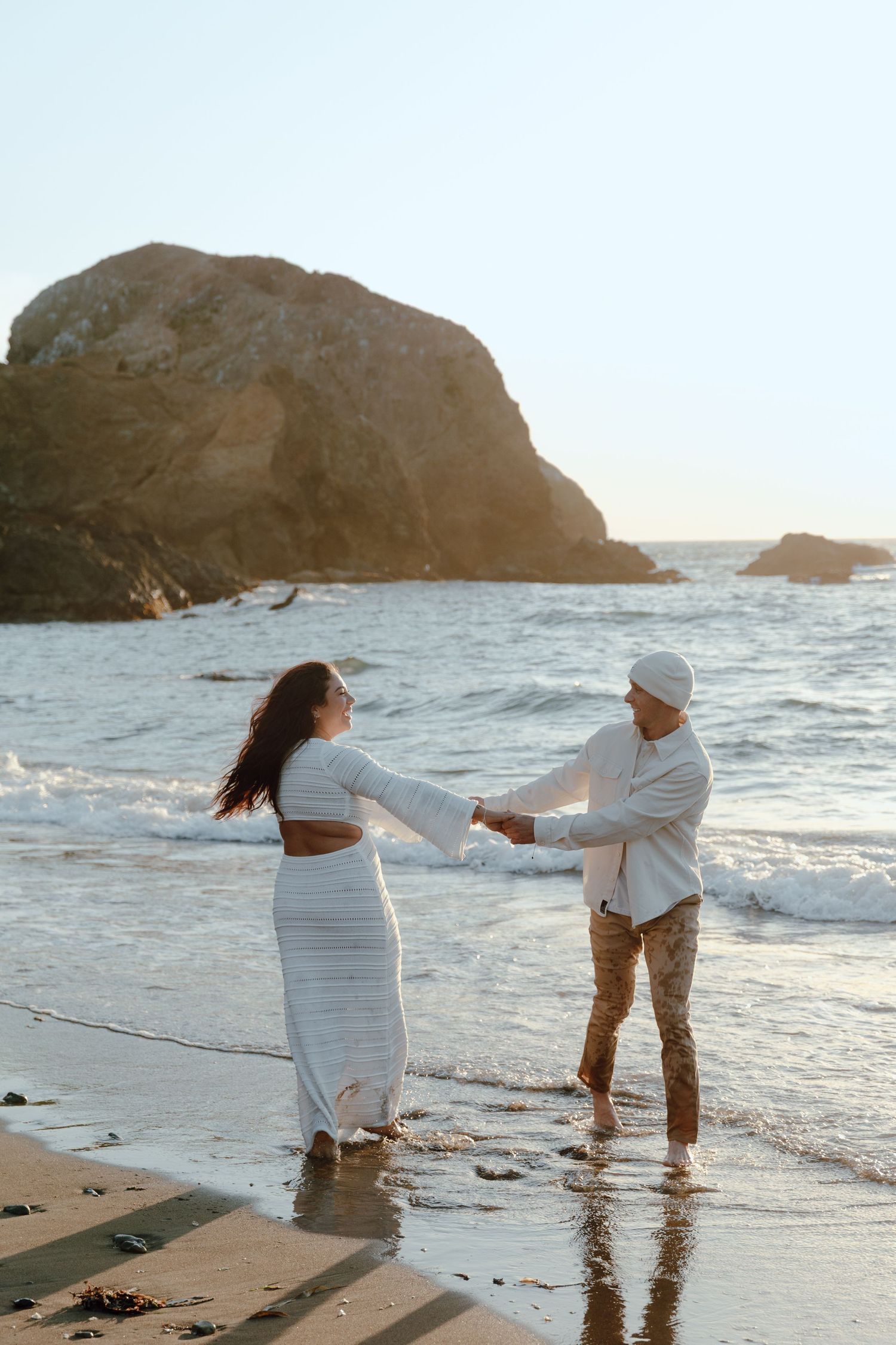 Couple holding hands and dancing together on a beach at sunset with rocky cliffs in the background.