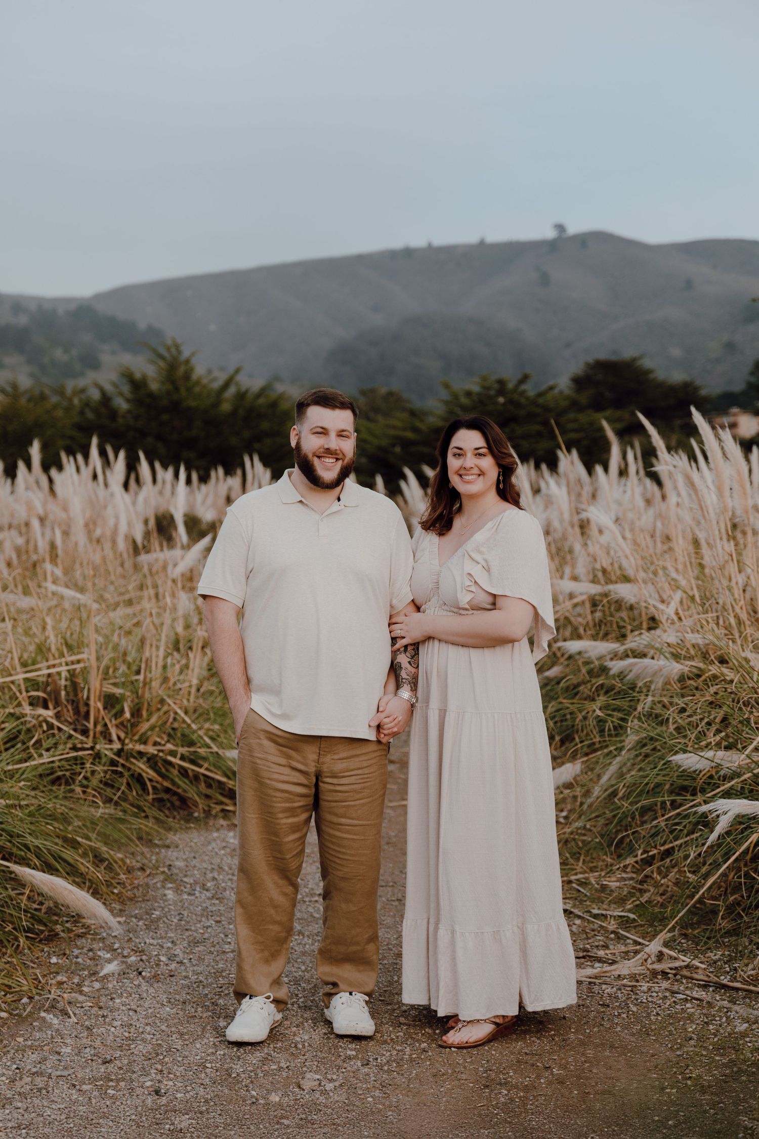 A couple stands together on a dirt path surrounded by tall pampas grass with rolling hills in the background at sunset.