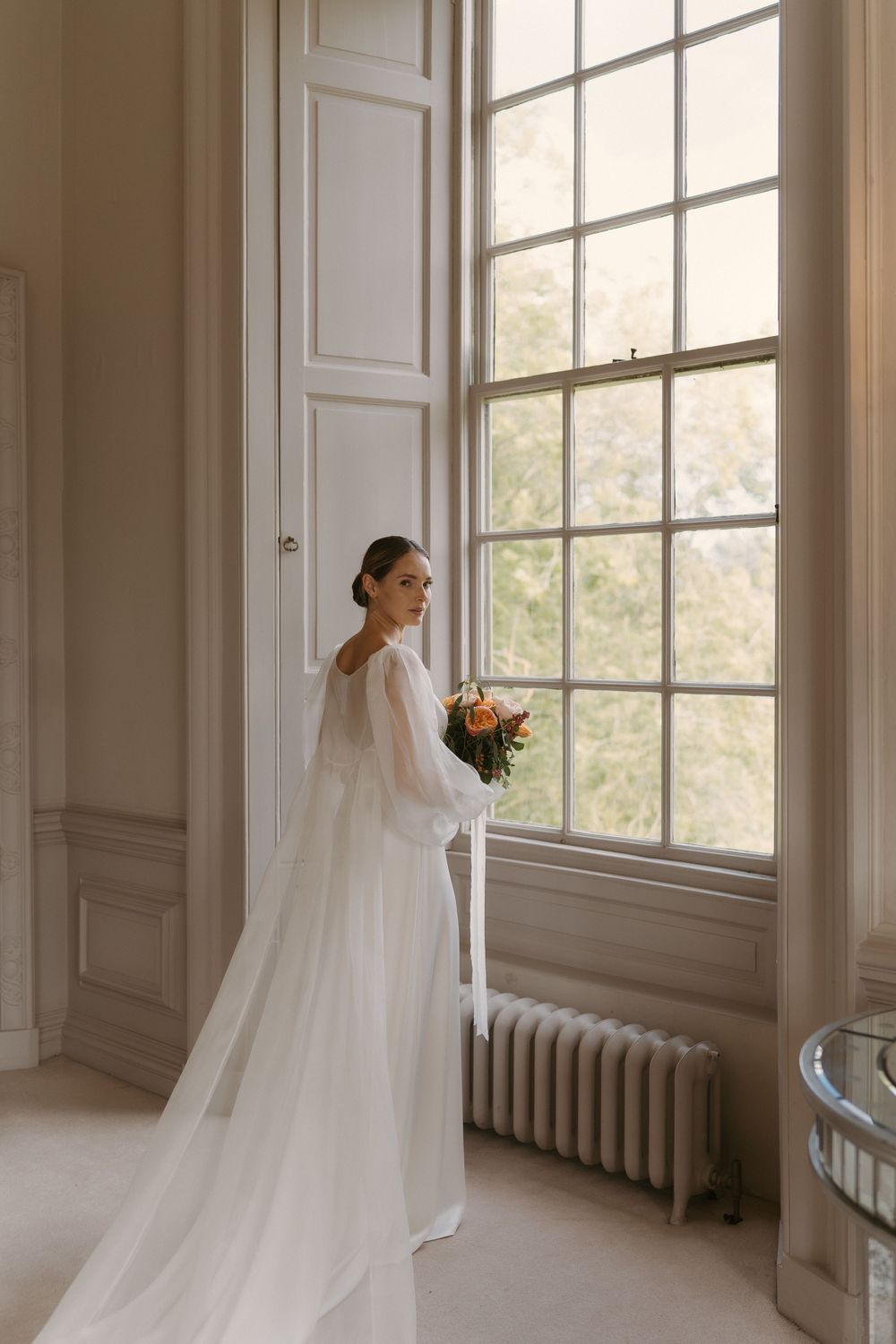 A person in an elegant white wedding dress and veil stands beside a tall window holding a bouquet.
