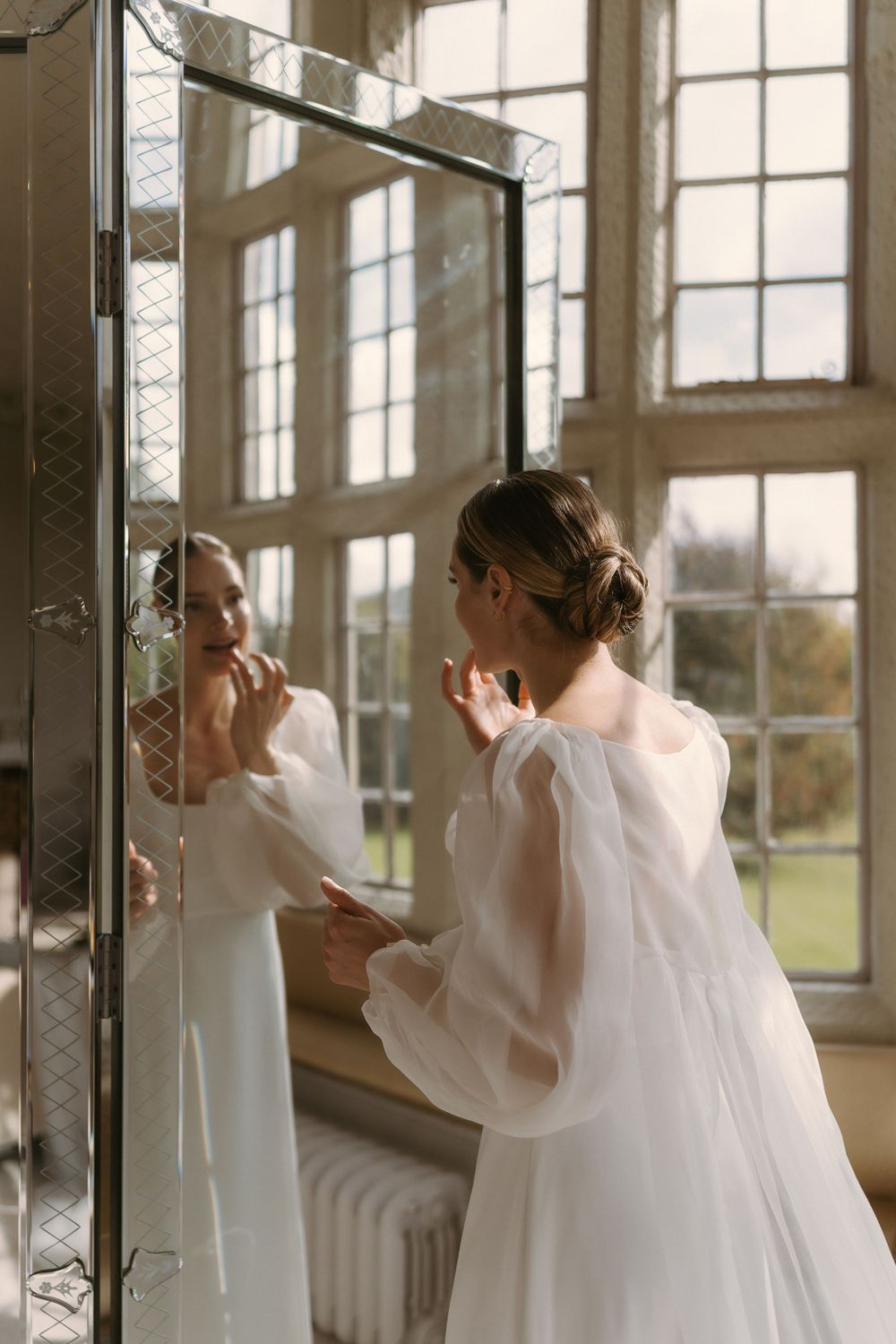 A person in a white wedding gown with sheer sleeves applies makeup while looking in a mirror in a sunlit room.