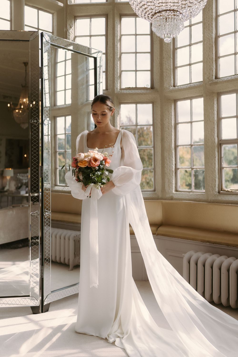 Elegant bridal portrait in sunlit room with crystal chandelier featuring long white dress and orange bouquet against tall windows.