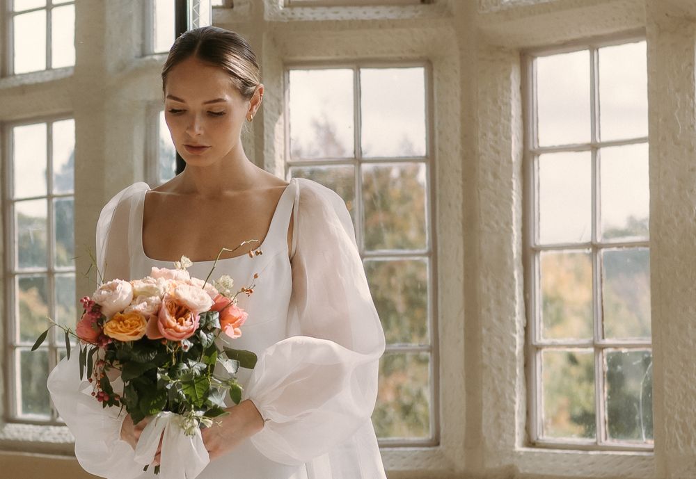 A bride in a white dress with puffy sleeves holds a bouquet of peach roses near sunlit windows.