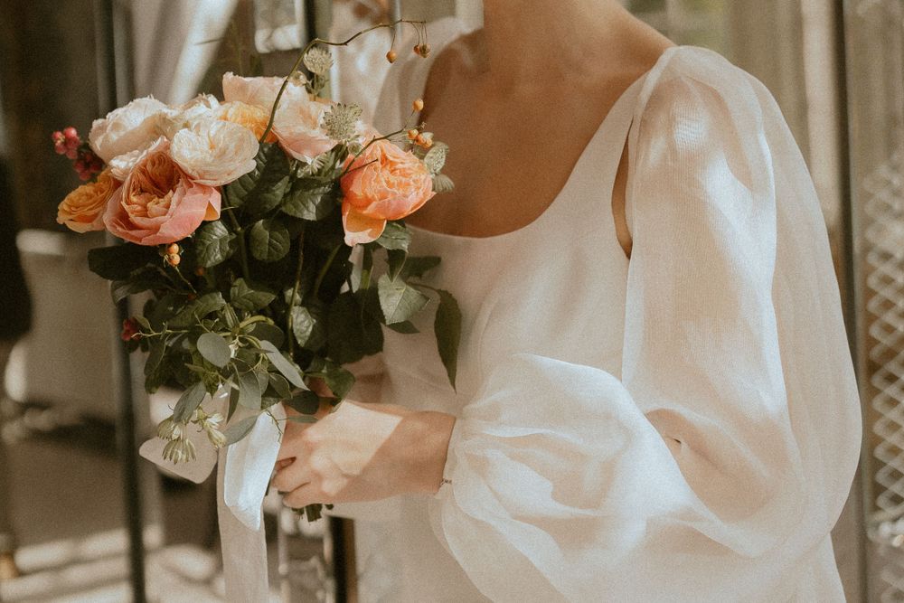A delicate bouquet of peach roses held against a flowy white dress with puffy sleeves in soft natural light.