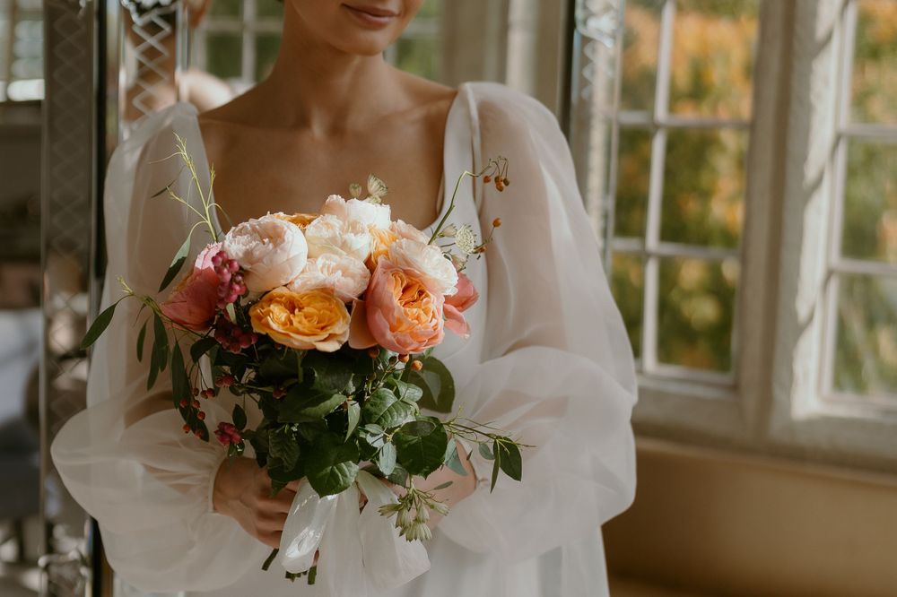 A soft pink and peach rose bridal bouquet held against a white flowing dress near sunlit windows.