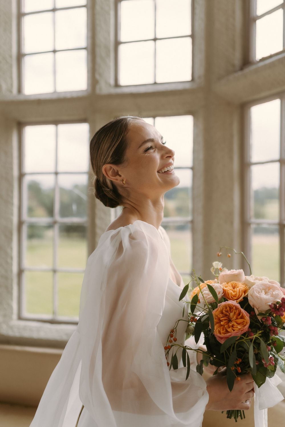 Elegant bride in minimalist long sleeve dress smiles joyfully while holding peach and coral floral bouquet by sunny window.
