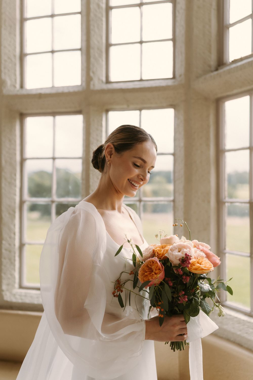 Radiant bride wearing classic white gown holds romantic peach and coral bouquet near bright window in natural light.