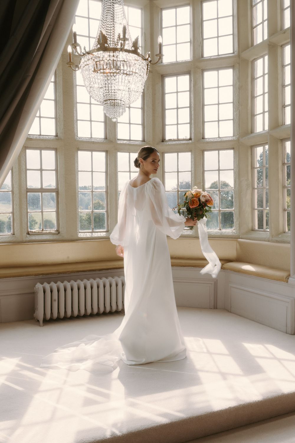 An elegant wedding dress with long sleeves in a bright sunlit room with a crystal chandelier and large bay windows.