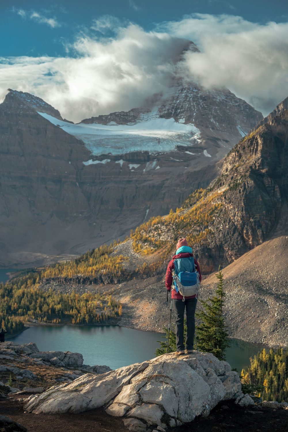 Hiker stands on rocky outcrop overlooking alpine lake and snow-capped peaks.