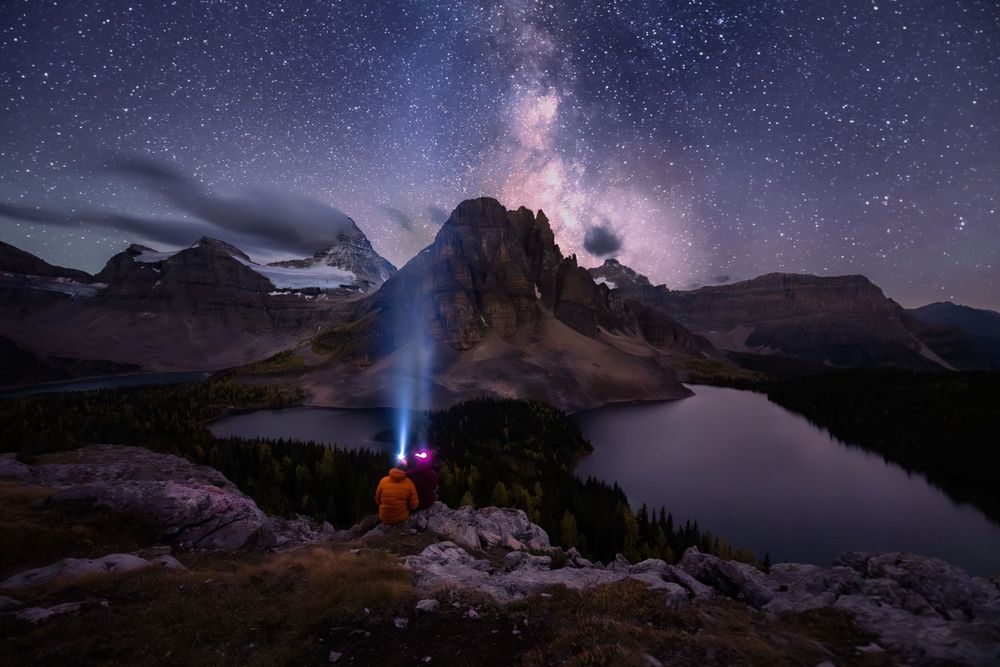 Milky Way galaxy over mountain peaks with flashlight beam shining from rocky viewpoint.