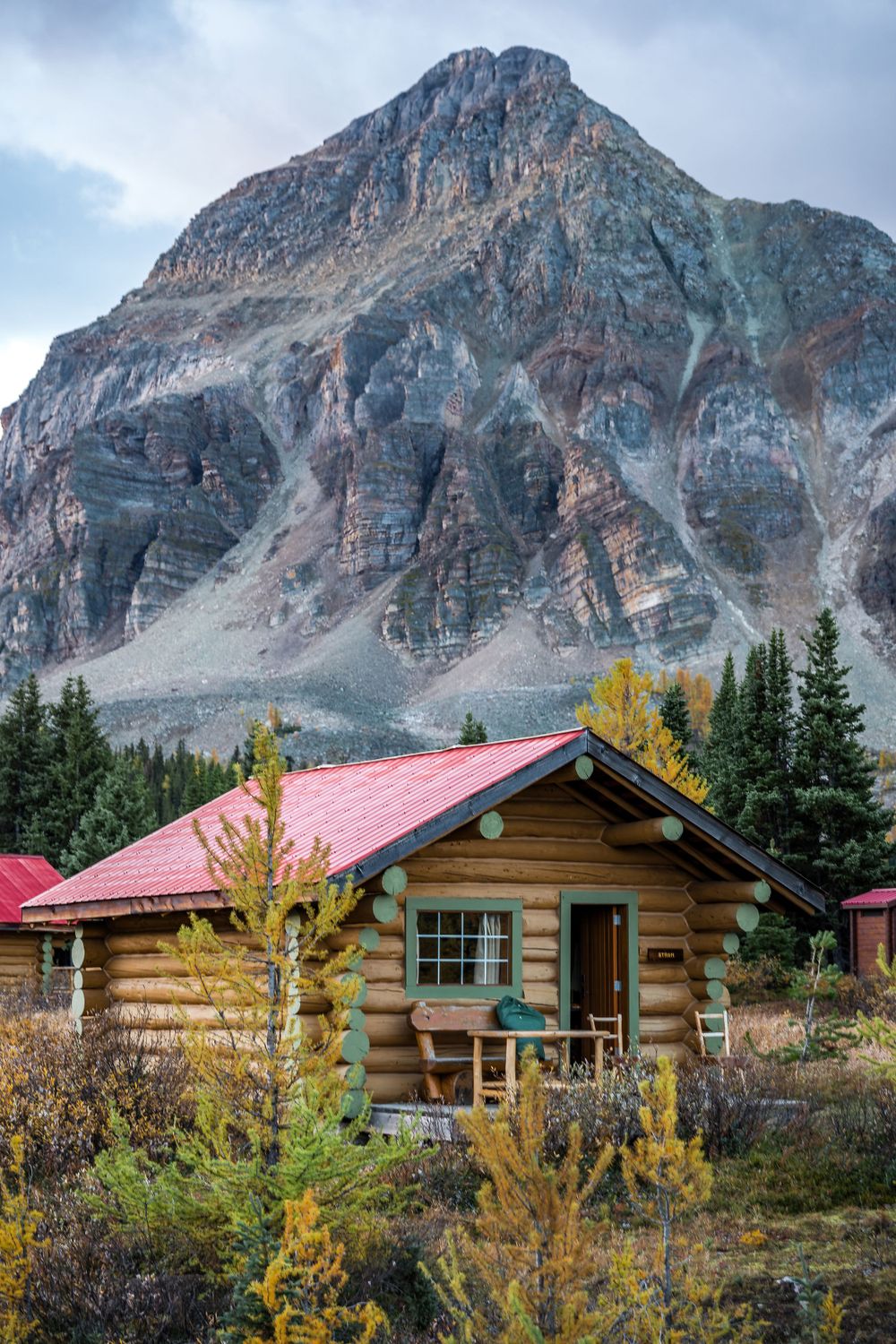 Rustic log cabin with red roof beneath towering mountain peak in autumn landscape.