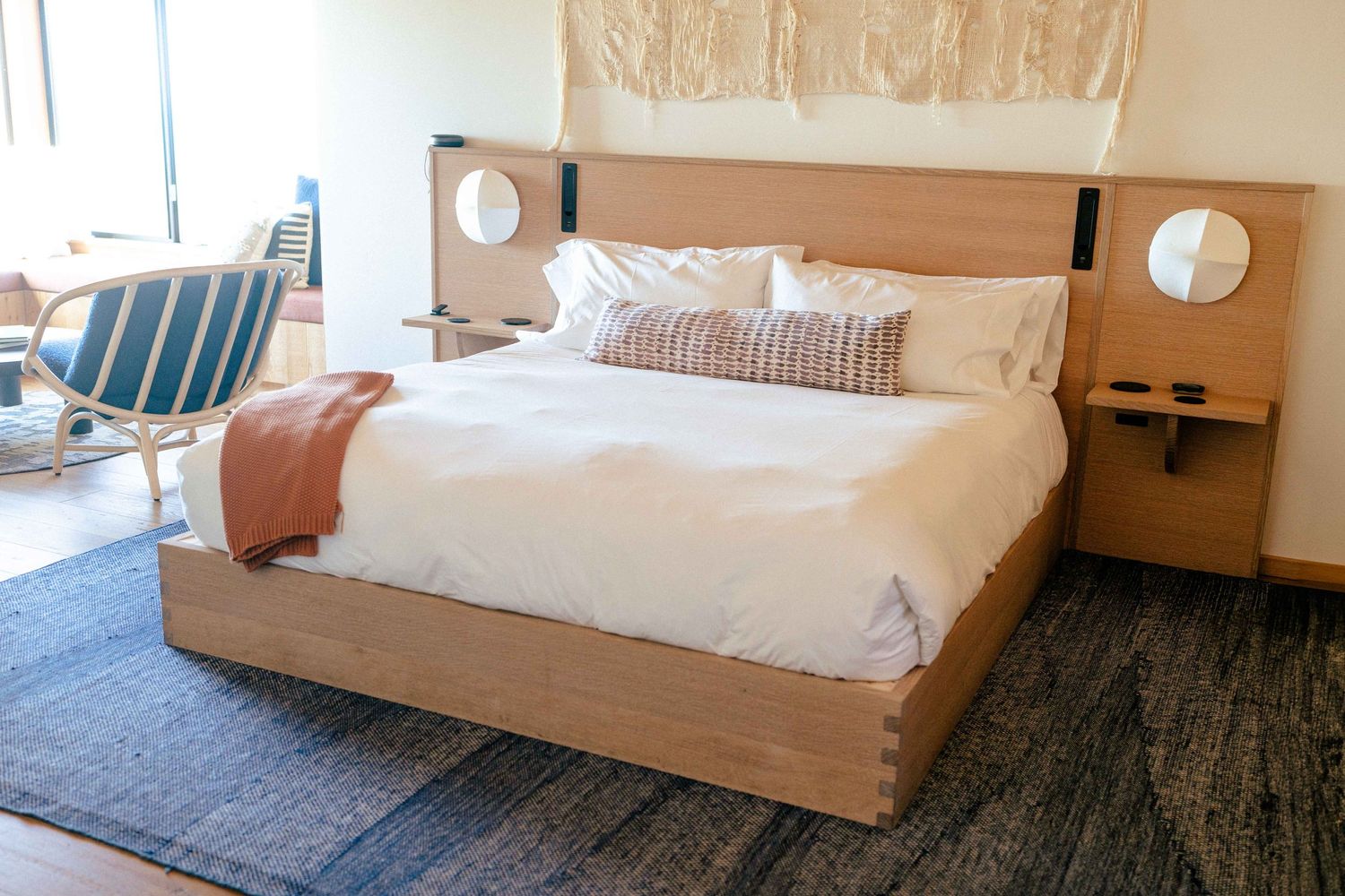 Modern Sea Ranch Lodge room interior with light wood platform bed, white bedding, built-in nightstands and blue accent chair.