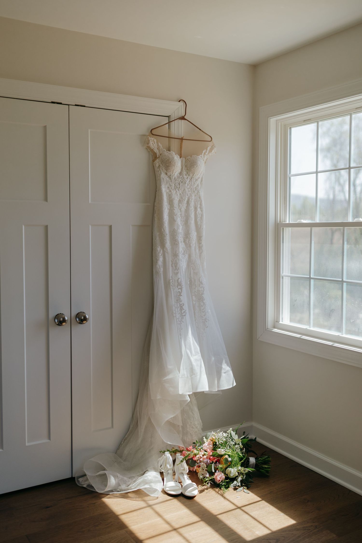 White lace wedding dress hanging in a sunlit room with bridal shoes and floral bouquet on hardwood floor.