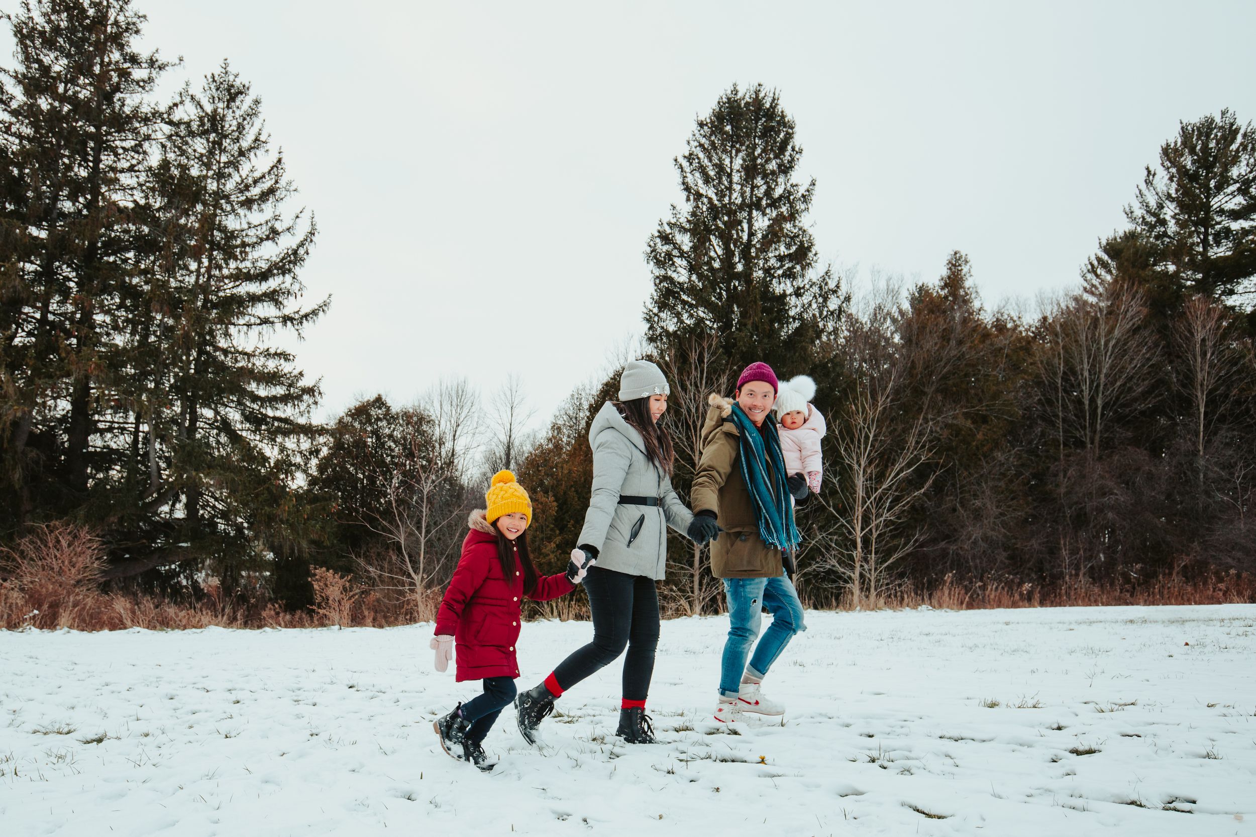 A joyful family bundled in winter coats plays together in a snowy field surrounded by evergreen trees.