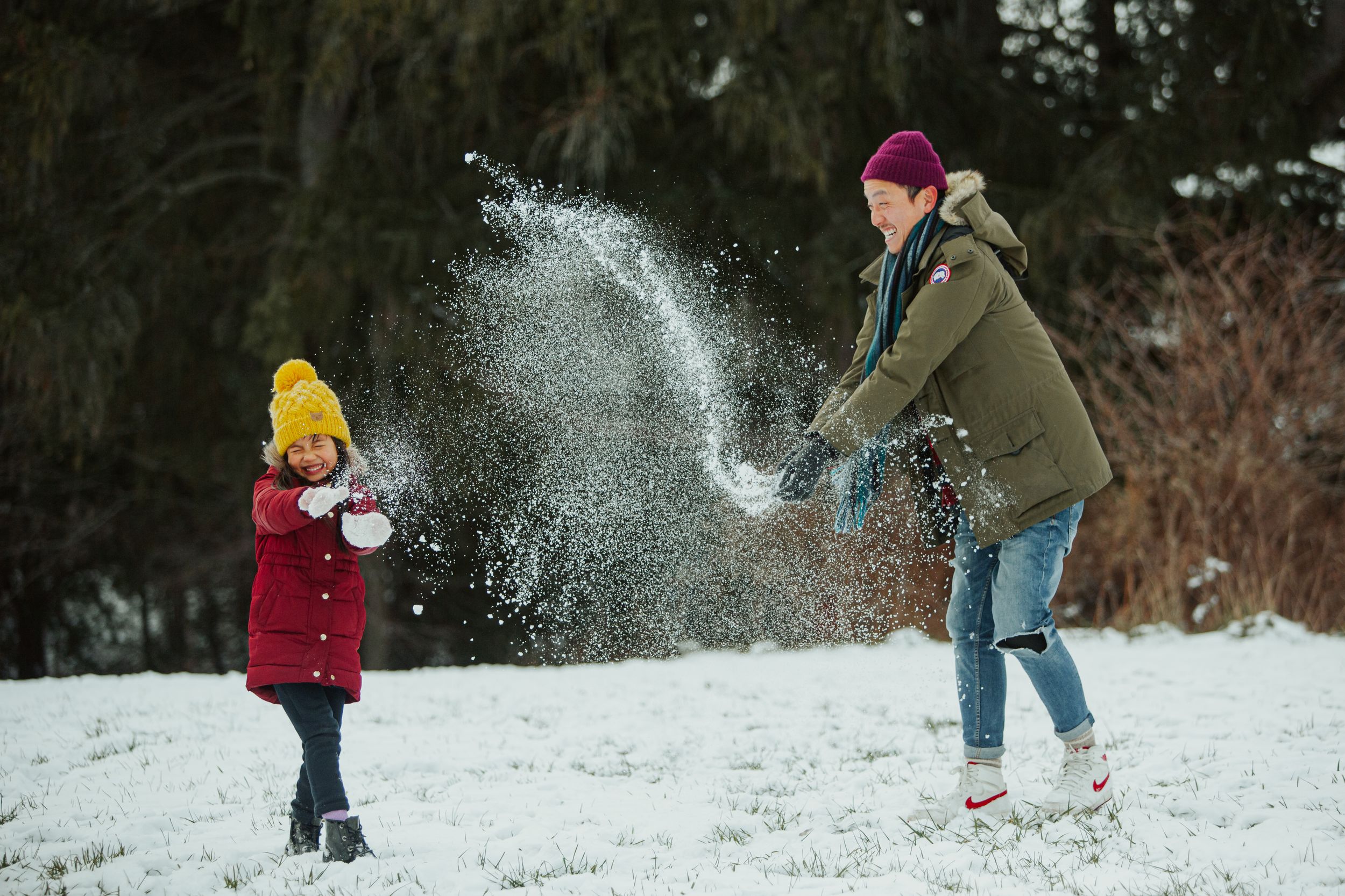 Two people playing in the snow with one throwing powdery snow while wearing winter coats.