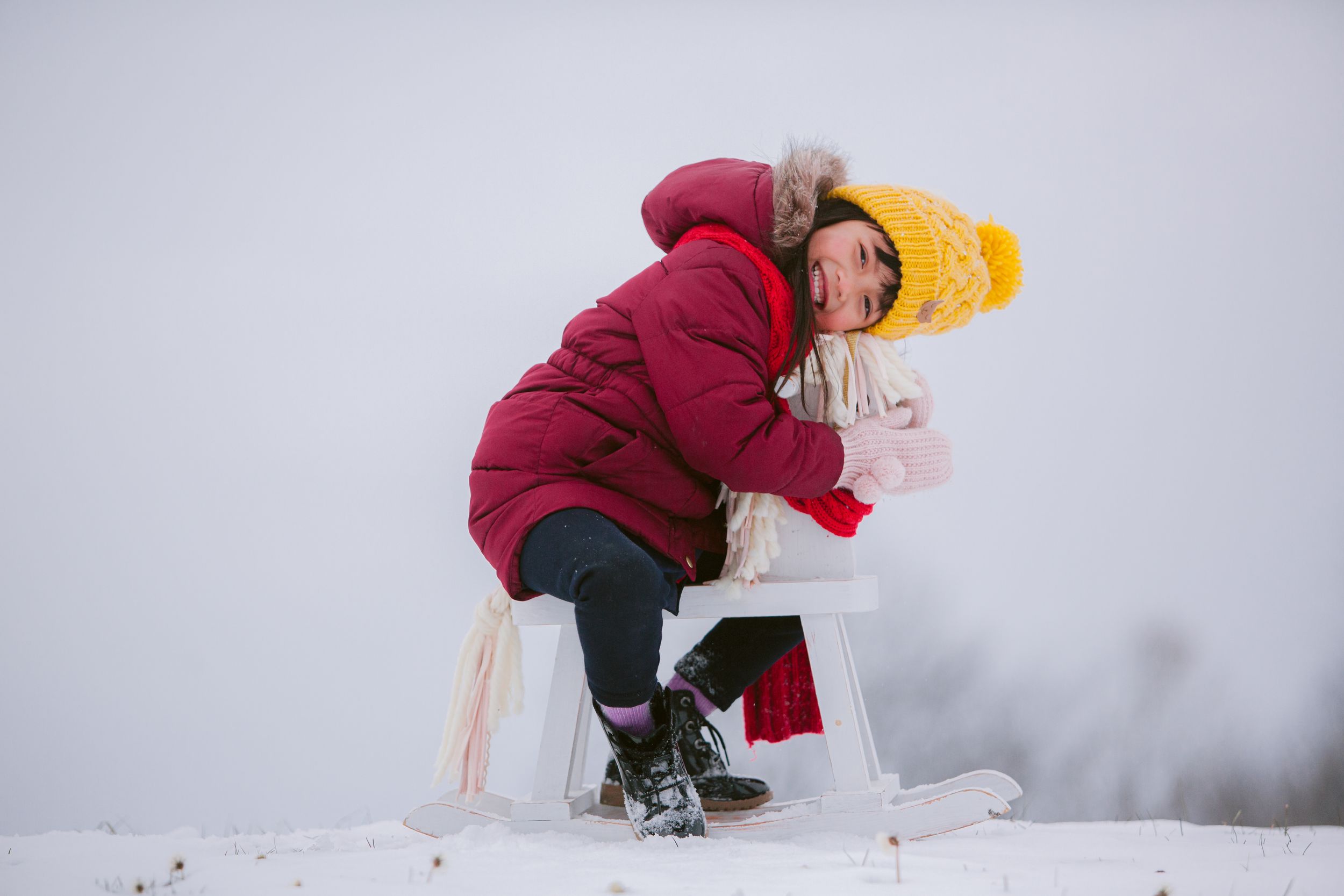 A winter scene shows someone in a red coat and yellow hat resting on a white stool in snowy conditions.