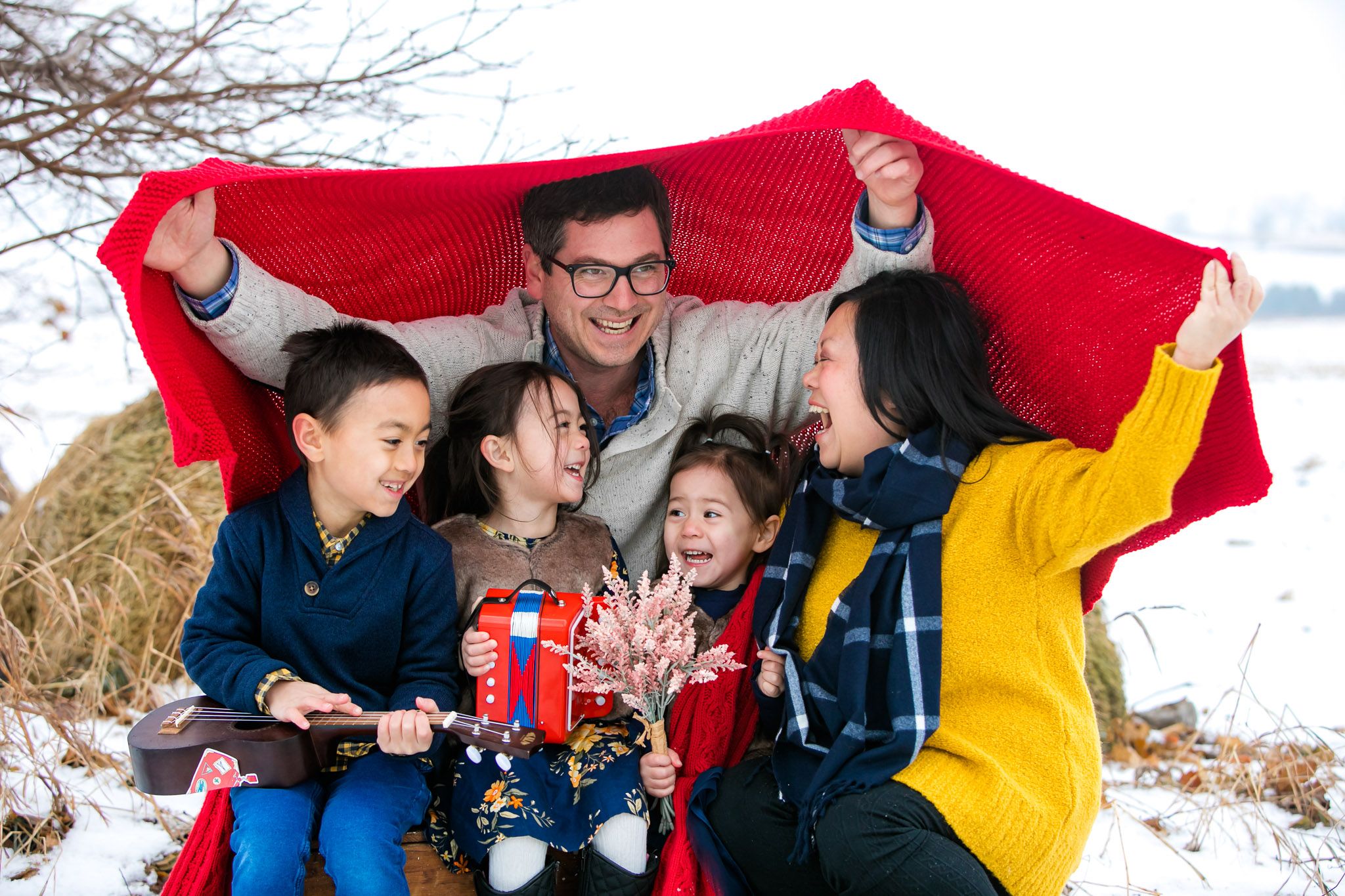 A family huddles together under a bright red blanket outdoors on a winter day, sharing joyful laughter and smiles.