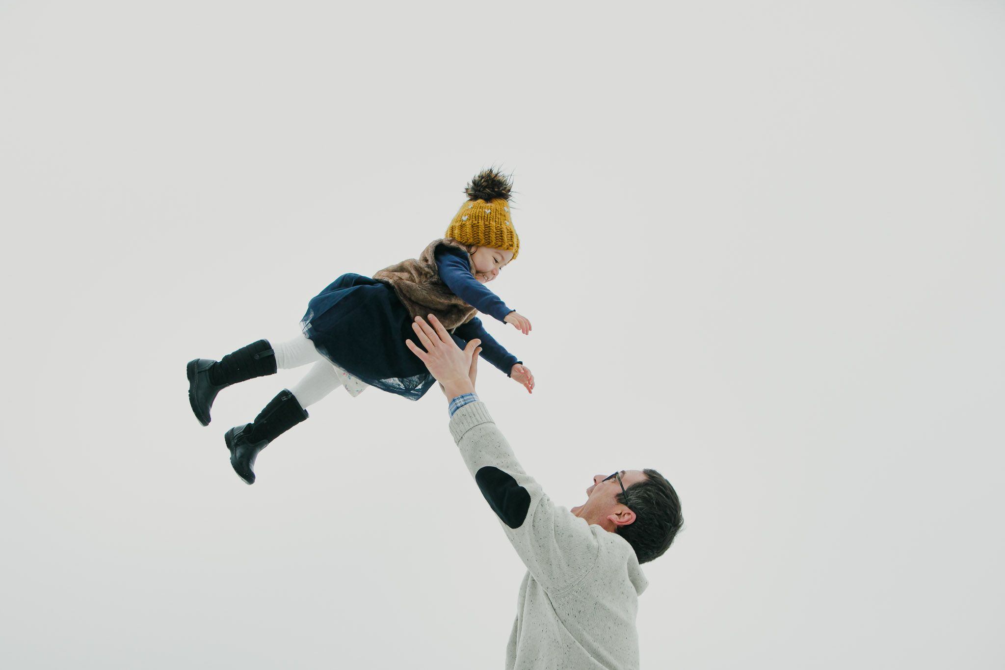 Parent in white sweater lifts child wearing yellow beanie and blue coat playfully against white background.