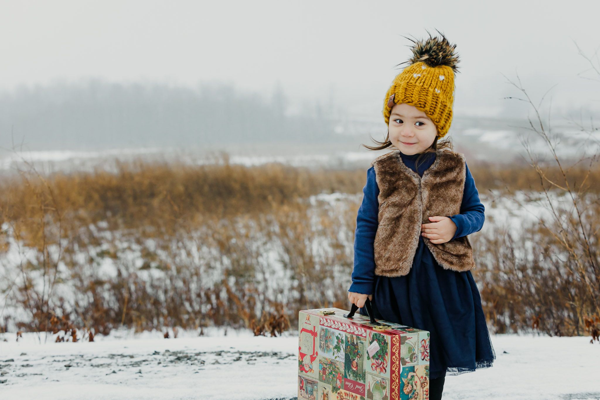Small figure wearing a yellow pom beanie and brown vest stands with vintage suitcase in snowy winter field.