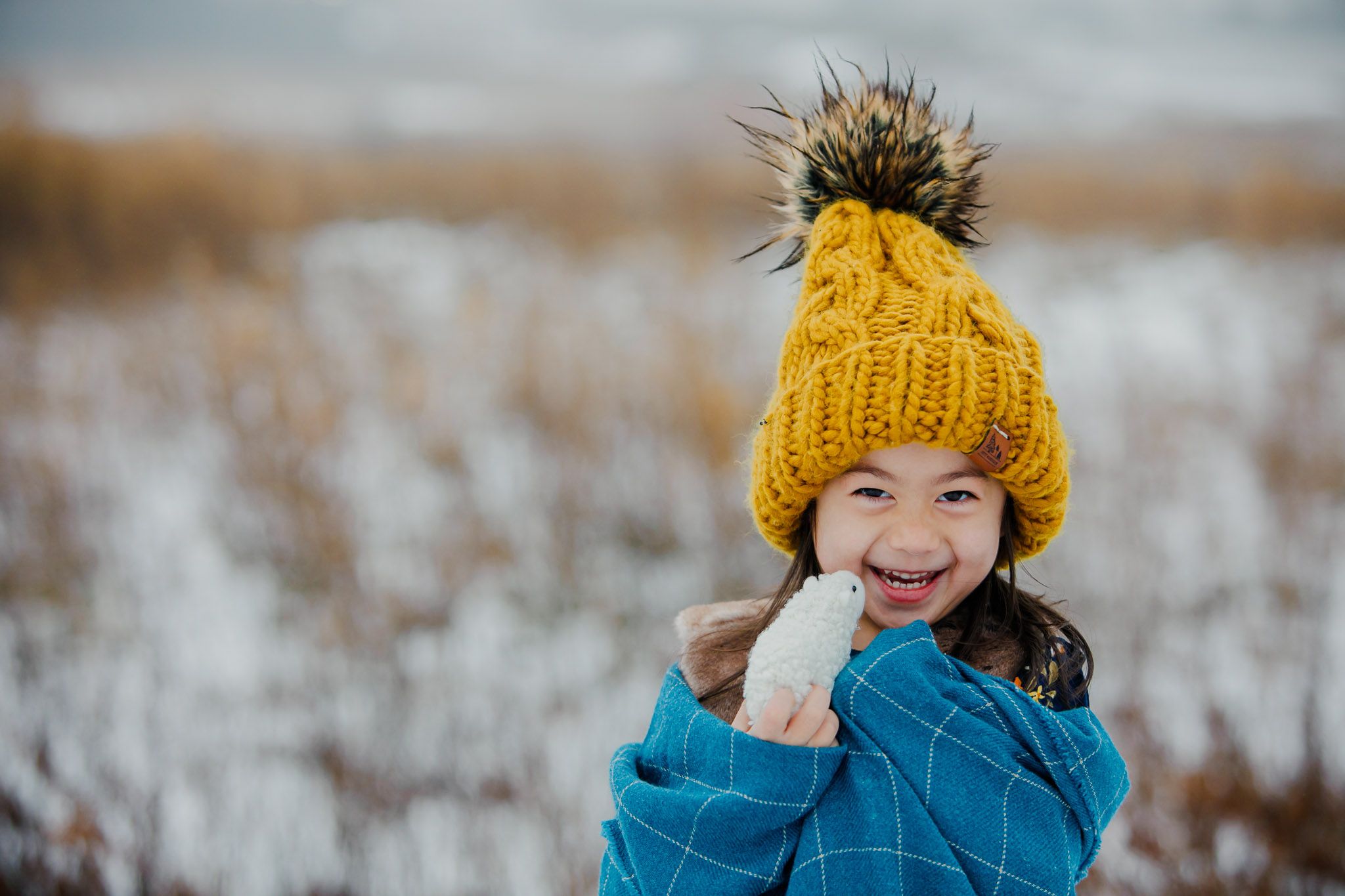 A cheerful person wearing a yellow knit pom beanie and blue winter coat smiles in a snowy field.