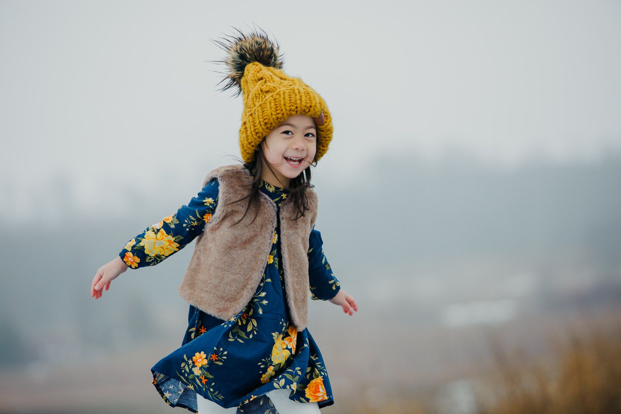 Child wearing yellow knit hat and floral dress plays outdoors on foggy day.