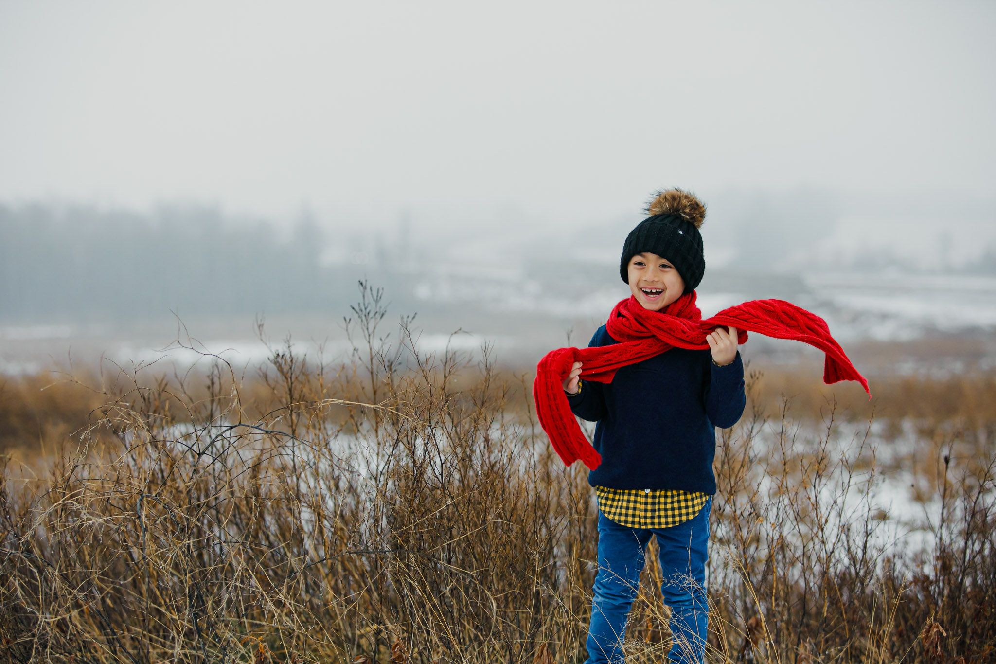 Child wearing winter clothing and red scarf playing outdoors in a snowy field with tall grass.