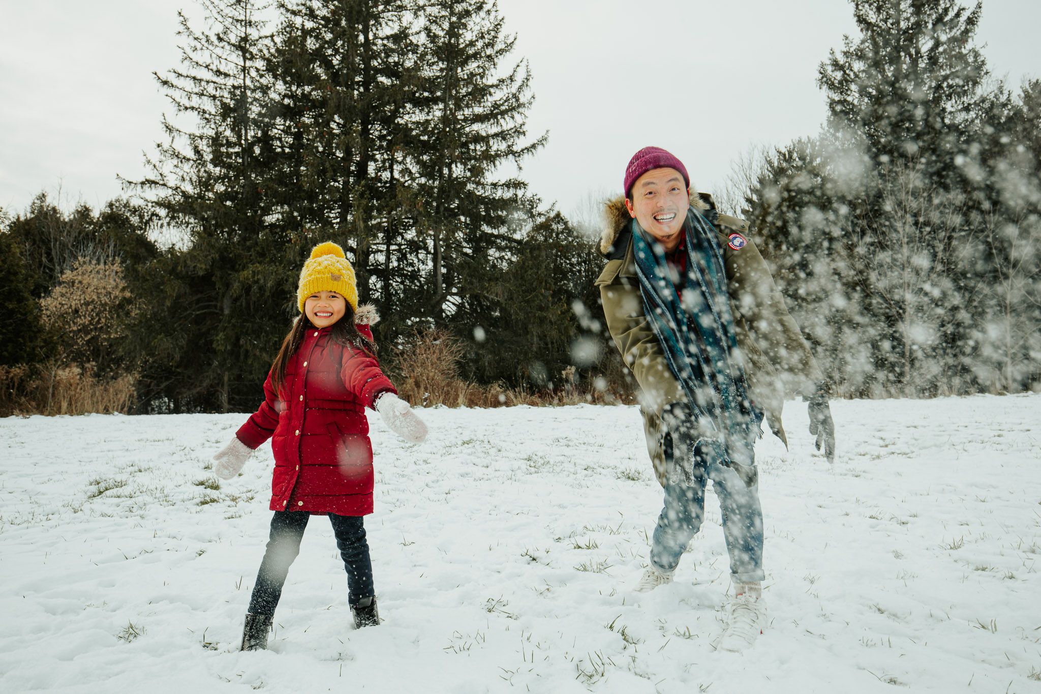Two people play in the snow, one wearing a red coat and yellow hat, the other in camouflage with a burgundy hat.
