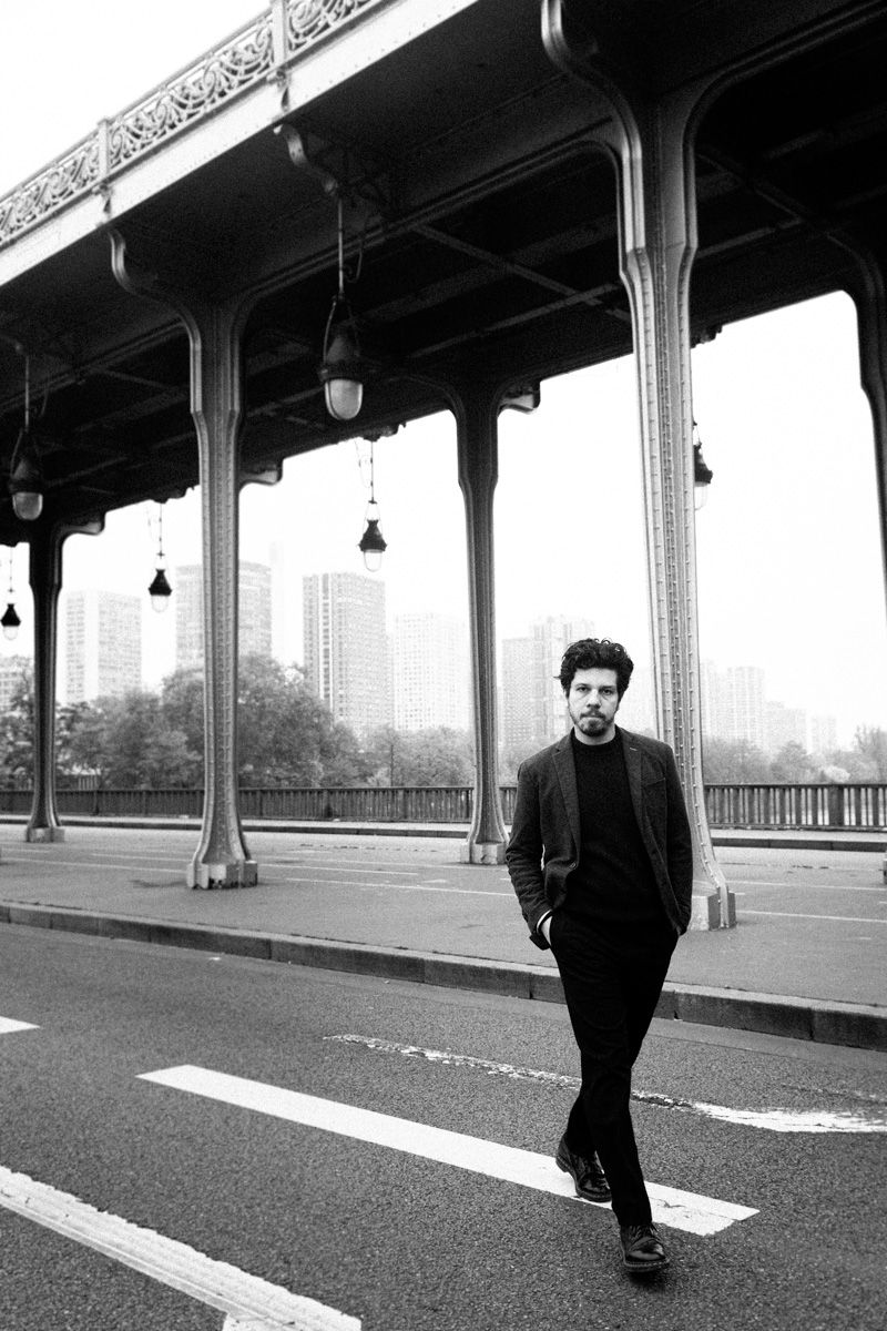 Black and white urban photograph of a person walking on a bridge with ornate metal architecture and city buildings in background.
