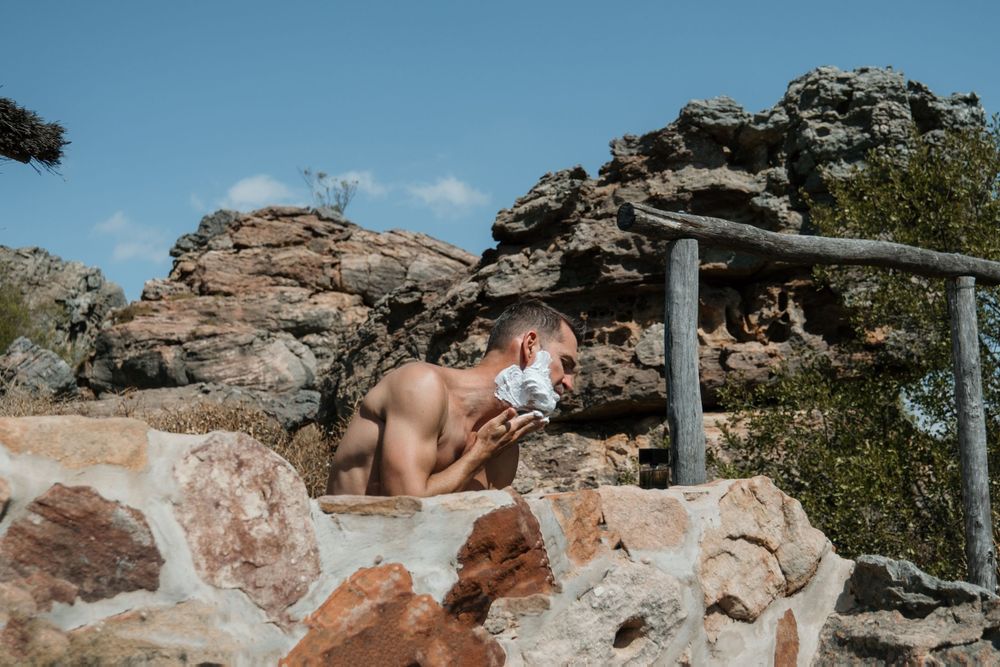 A shirtless person rests on craggy red rocks beneath a shelter during a sunny summer day.