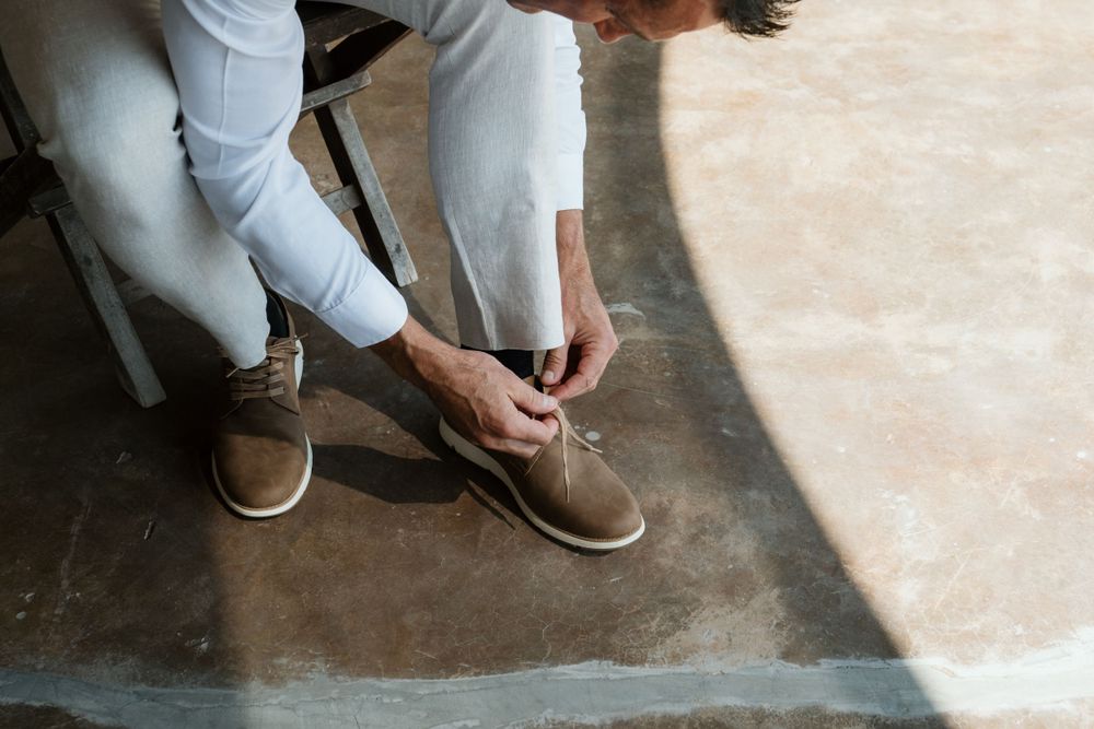 Someone wearing light colored pants ties the laces on brown suede shoes while sitting on a concrete floor.