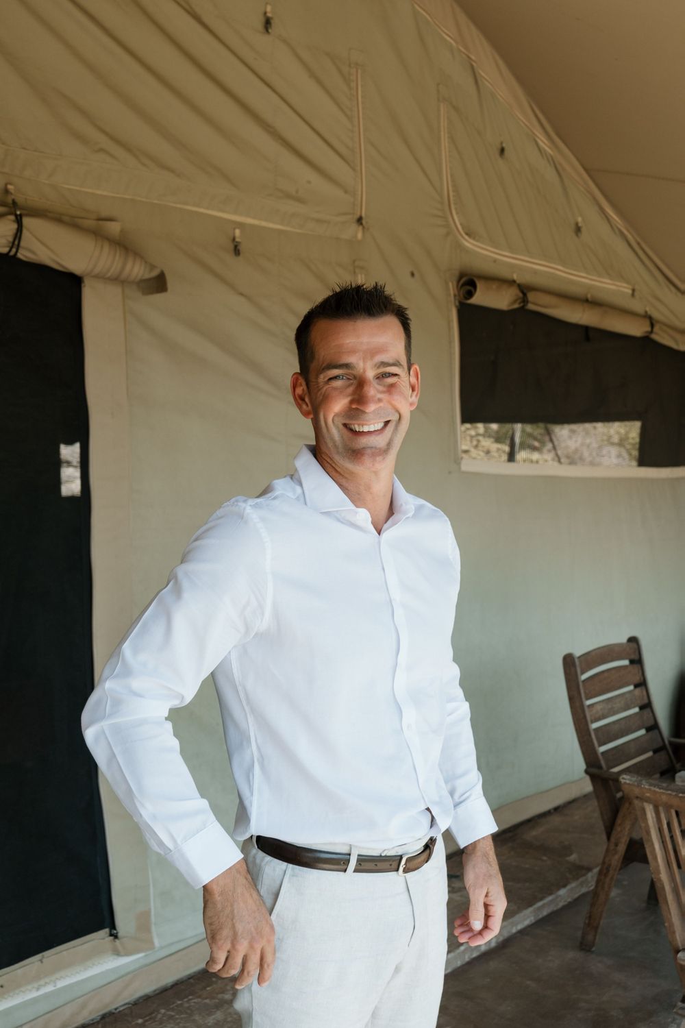 Someone wearing a crisp white button-down shirt and khaki pants smiles warmly while standing in a covered outdoor area.