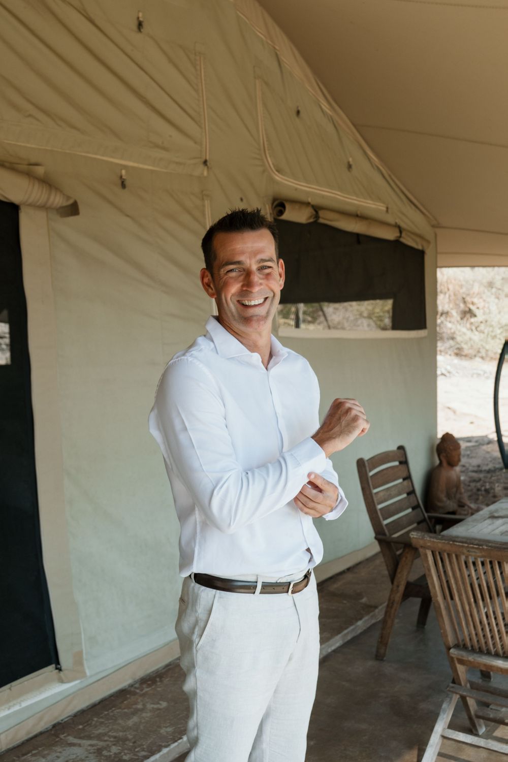Someone in an all-white outfit smiles while adjusting their sleeve on a covered outdoor porch with wooden furniture.