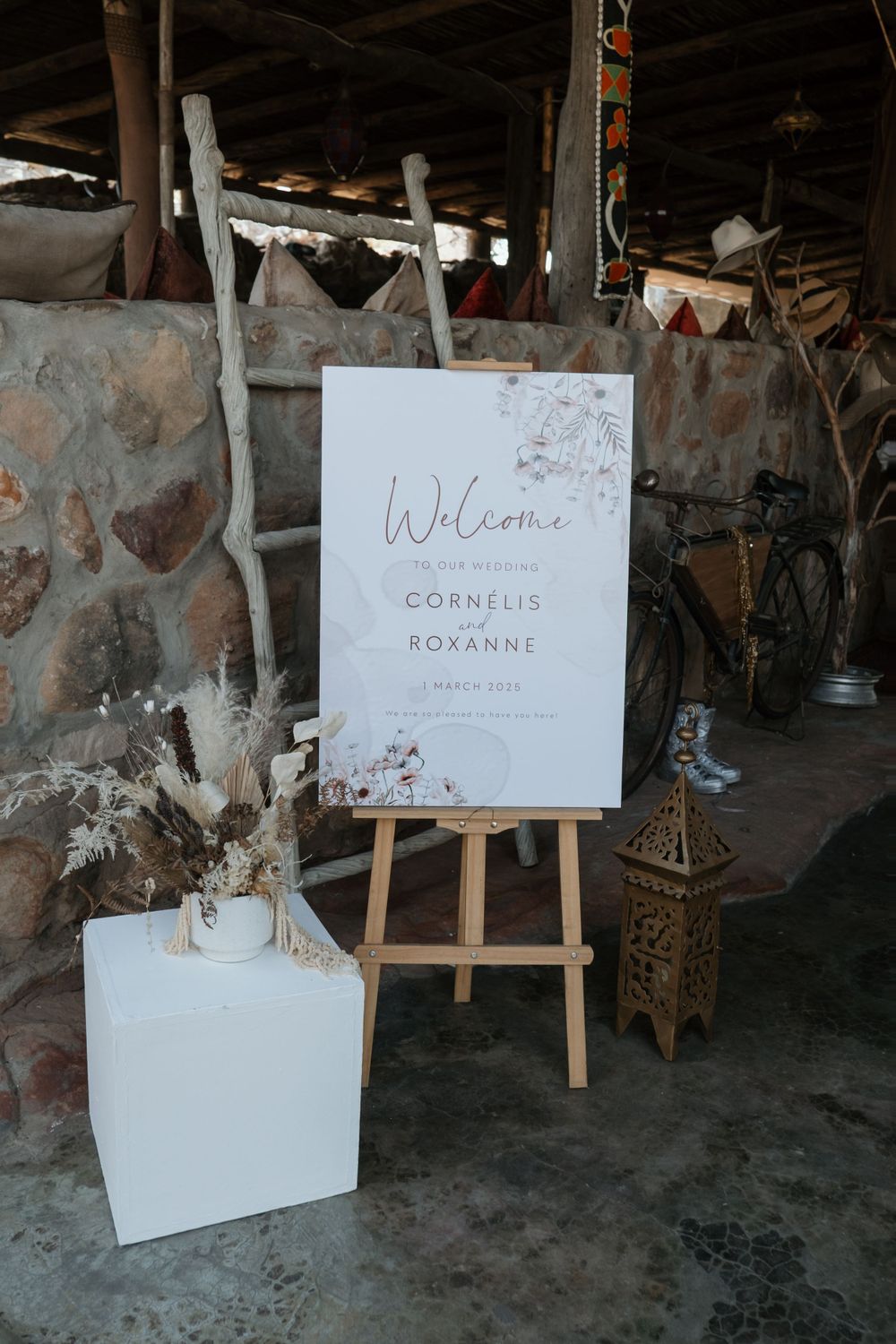 A rustic wedding welcome sign on a wooden easel with dried flowers and a white pedestal in a stone-walled venue.