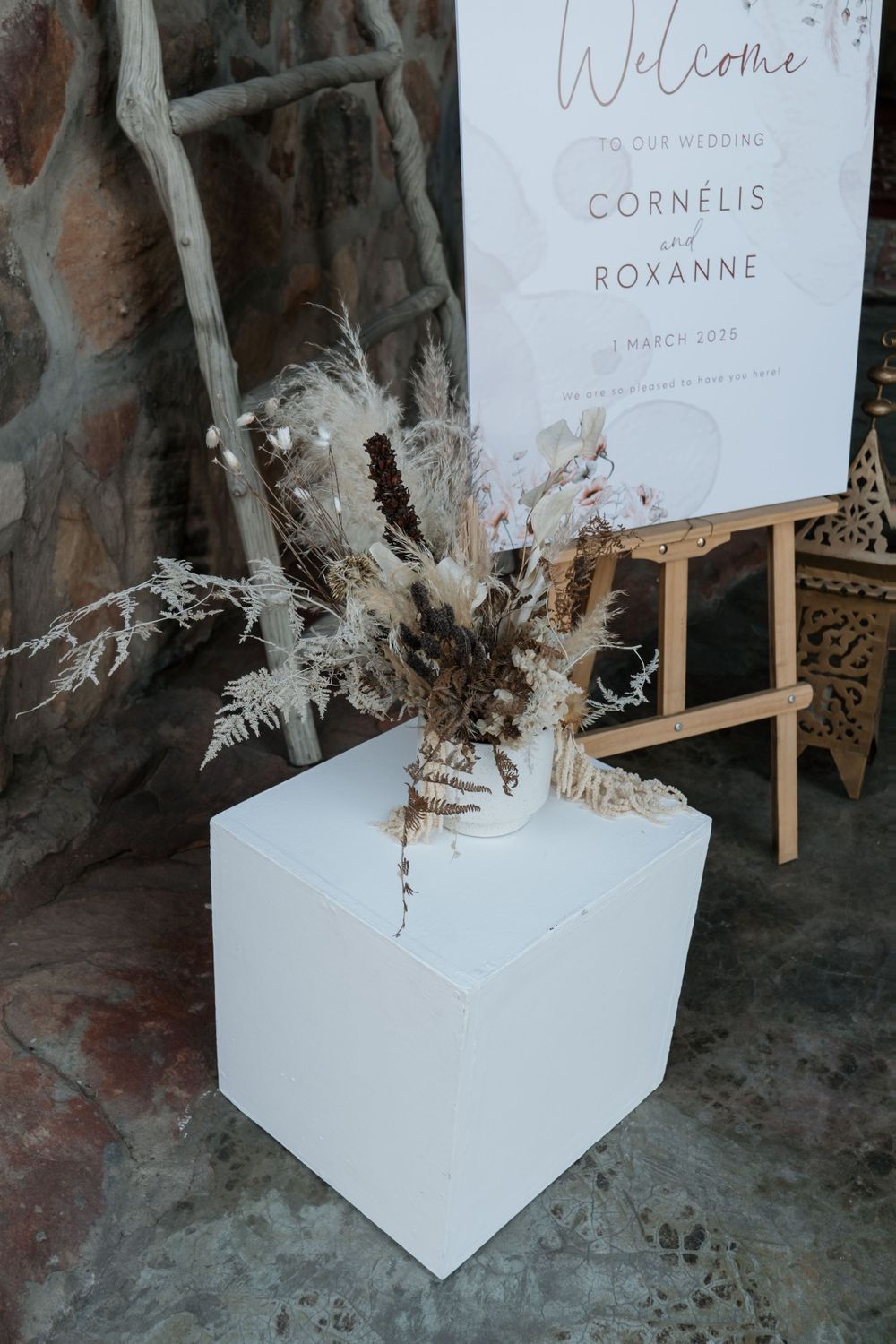 Wedding welcome sign and dried floral arrangement in white geometric vase against dark backdrop at venue entrance.