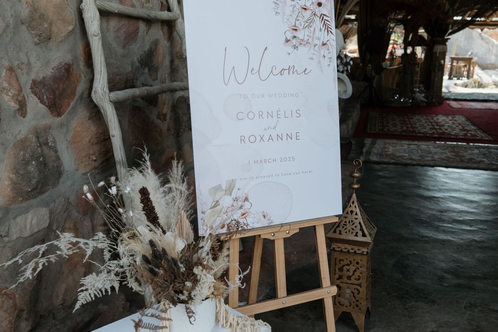 A wooden easel displays a welcome sign with dried floral decorations against a stone wall at a rustic outdoor venue.