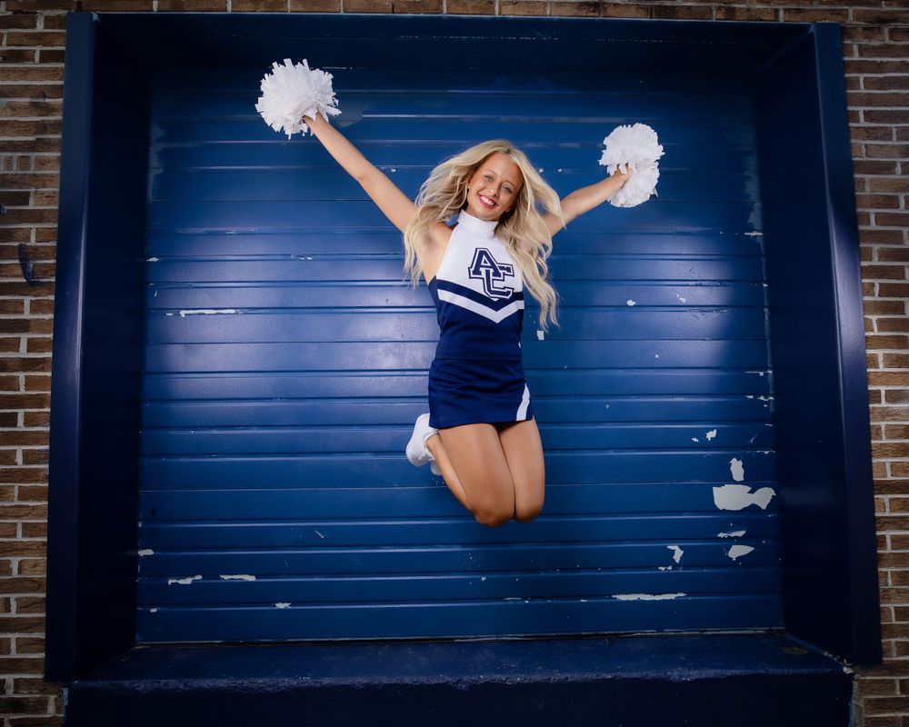A cheerleader in a navy and white uniform jumps high with pom-poms raised against a blue garage door background.