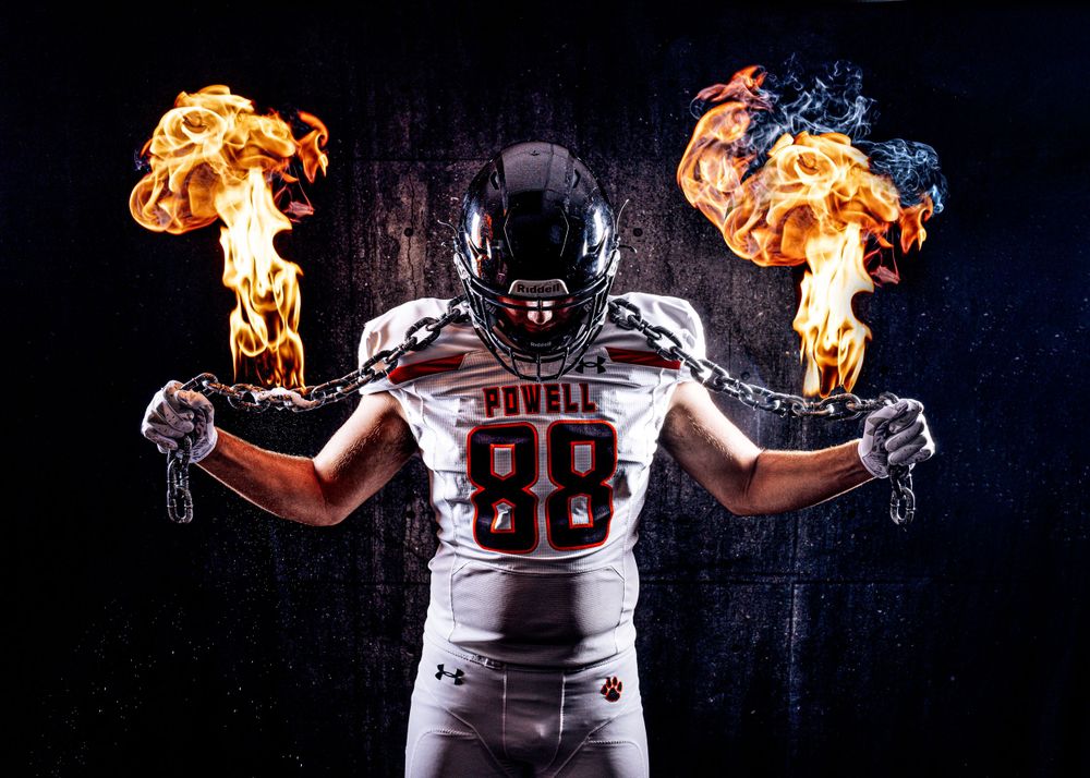Football player wearing white uniform stands with flaming torches in both hands against dark background.