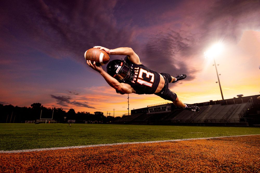 Football player diving for a catch in dramatic sunset lighting on a baseball field with vibrant orange sky.